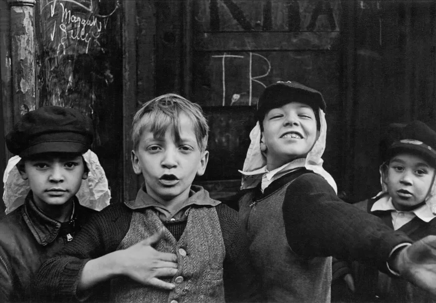 Black-and-white documentary photograph of four boys in flat caps and knitted sweaters crowded in front of a weathered wooden storefront, with chalk writing on the boards behind them.